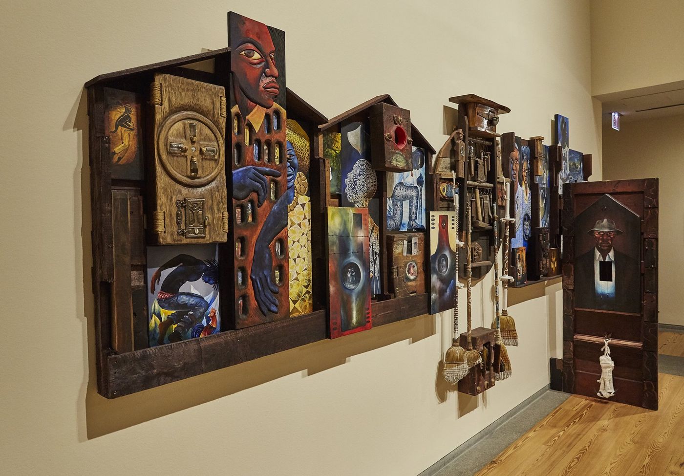 A Distant Holla: a long wall of house-shaped assemblages in carved and painted wood, with figures, doors, brooms and painted panels, installed in a museum gallery