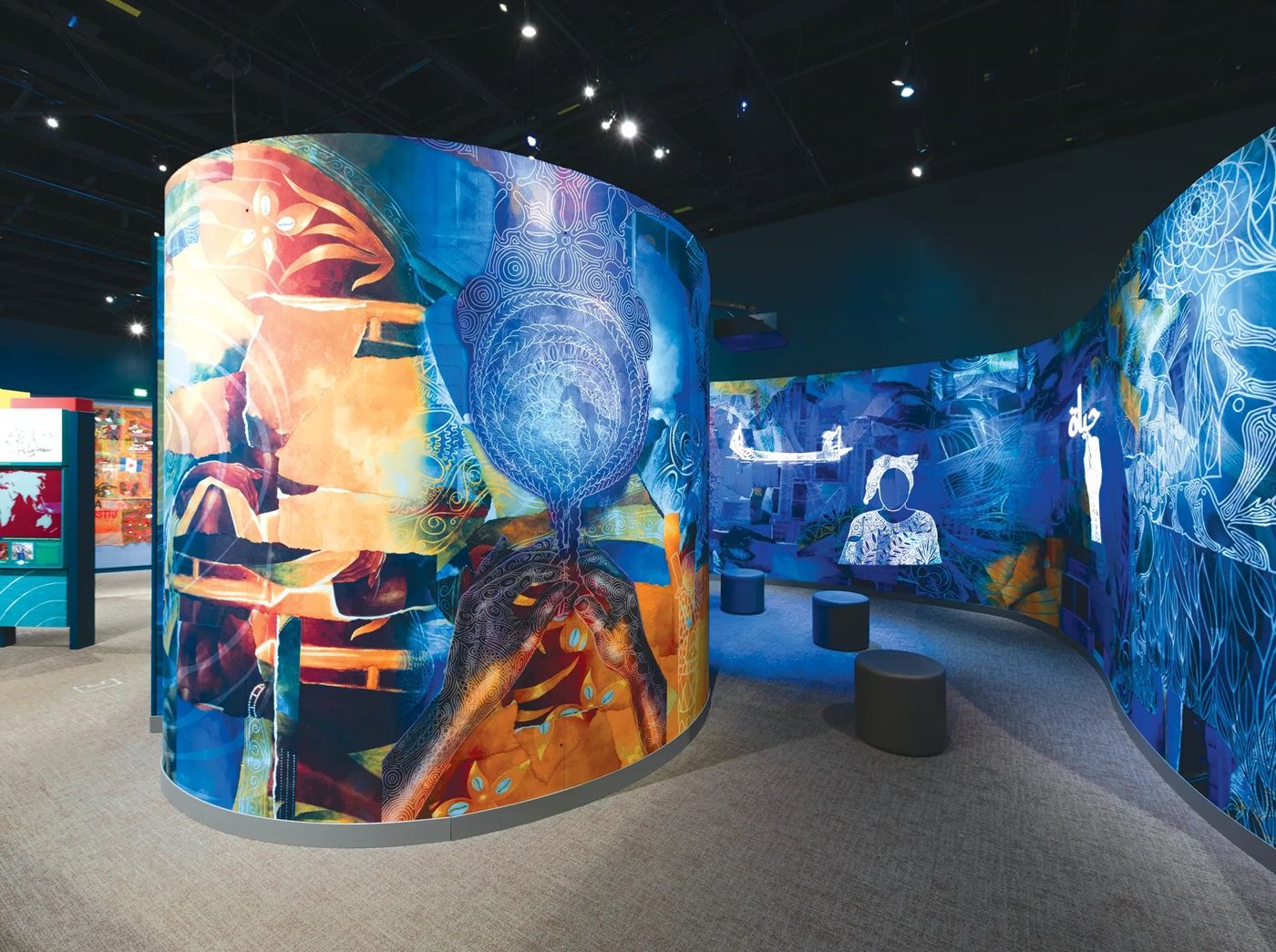 Universe of Freedom Making at the Smithsonian NMAAHC: a curved freestanding wall and a long sweeping wall printed with layered collage in orange and deep blue, with patterned hands, faces and a boat, and low seats in a lit alcove