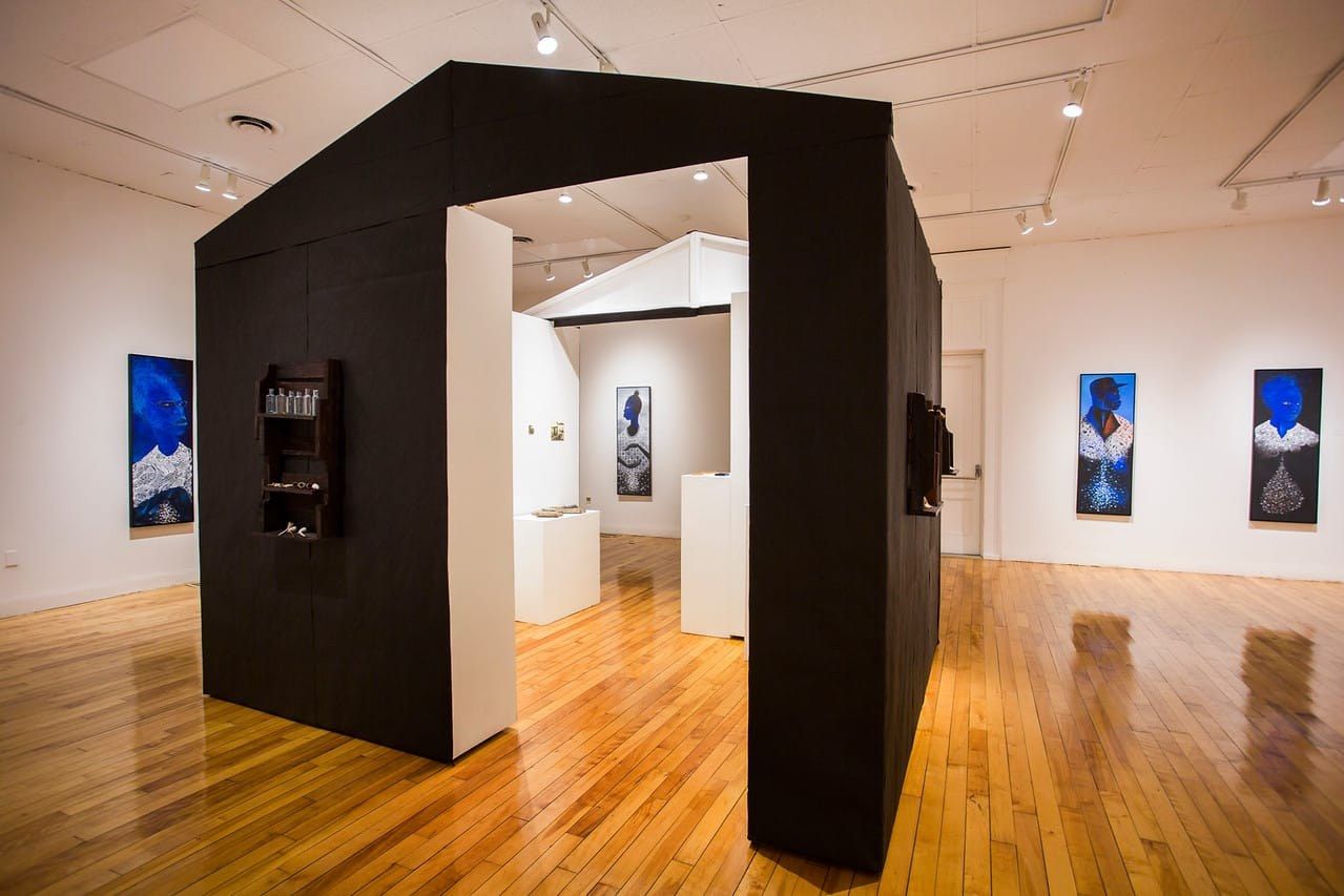 A black house-shaped structure with open doorways stands in a gallery with wood floors, a dark shelf of small bottles on its side; tall blue portrait paintings hang on the white walls around it