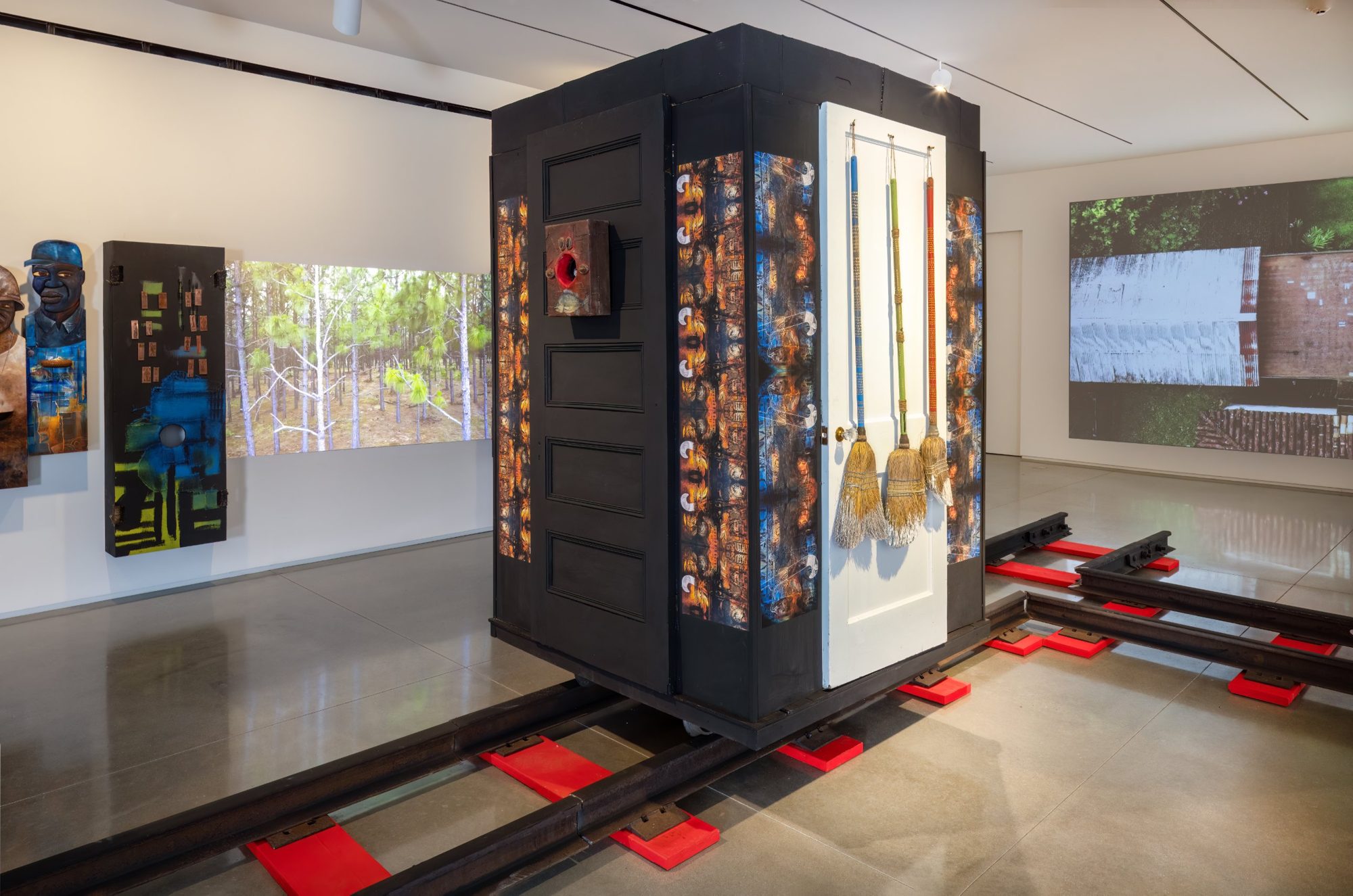 A black cabinet of doors with three hanging brooms, set on railroad tracks, with projected forest footage behind