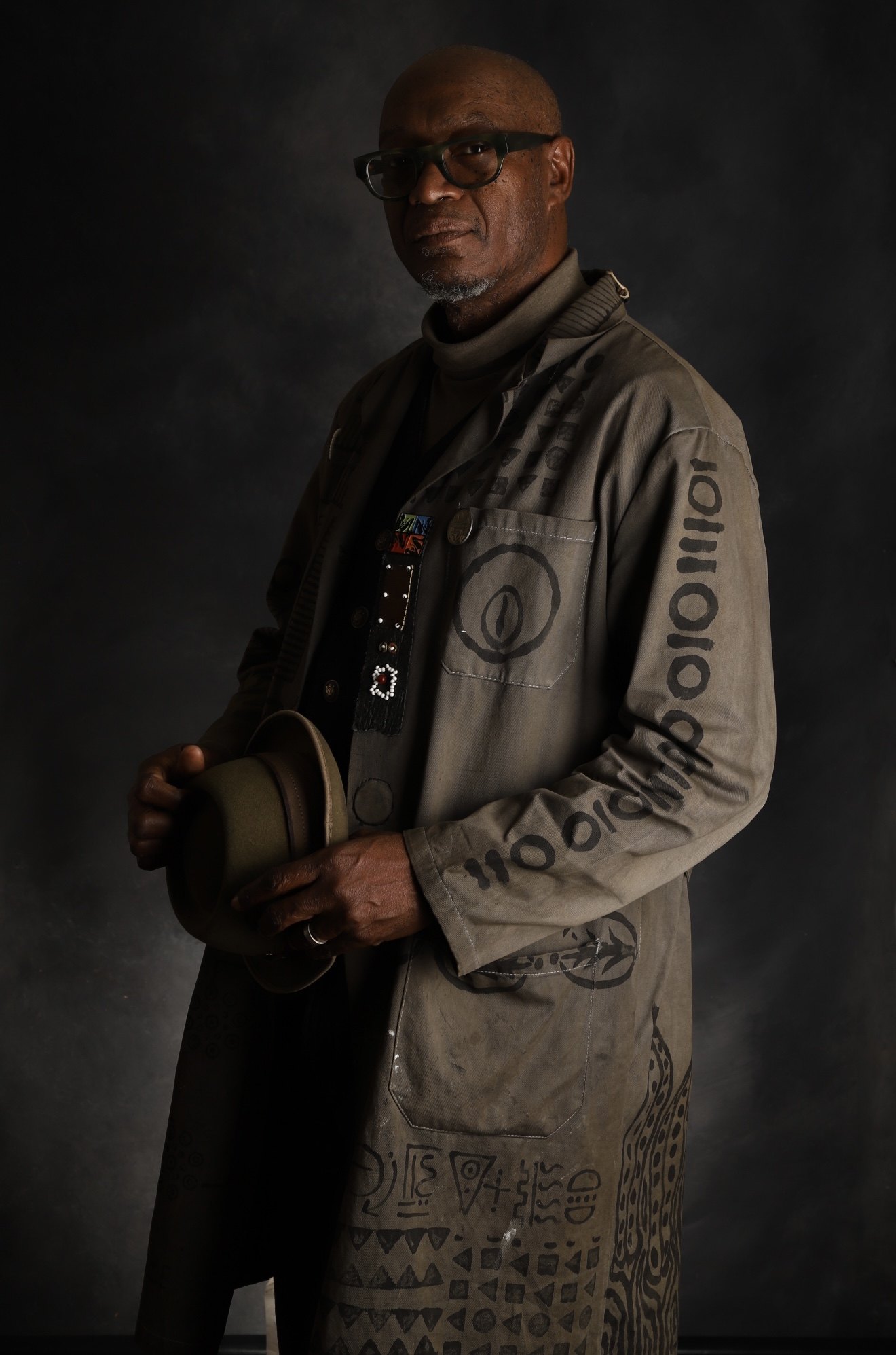 Portrait of Daniel Minter in a studio coat hand-painted with symbols, holding his hat, against a dark background