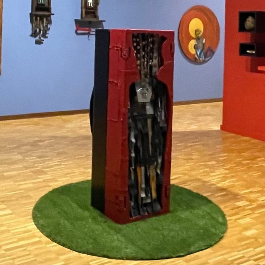 Red and black standing sculpture with a carved figure inside, on a green circle, in the Thresholds installation at the Haggerty Museum of Art